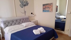 A neatly made bed with a blue bedspread and white pillows, a bedside table with a clock, a wall art of a flower, and a painting of a coastal scene. - Hotel Suisse Genova (Genoa)