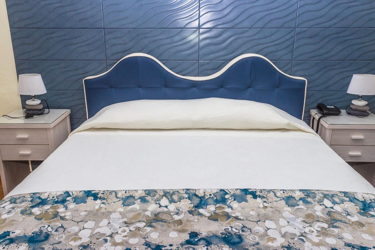 Una cama con cabecera azul, ropa de cama blanca y una manta con estampado floral azul. Hay dos mesitas de noche, cada una con una lámpara y un teléfono.