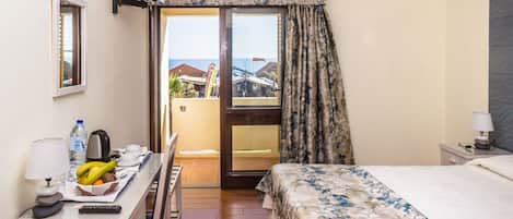 Una camera d'albergo con un letto, una scrivania con lampada, una sedia, un tavolino con frutta e un bollitore, e vista mare tramite una porta in vetro.