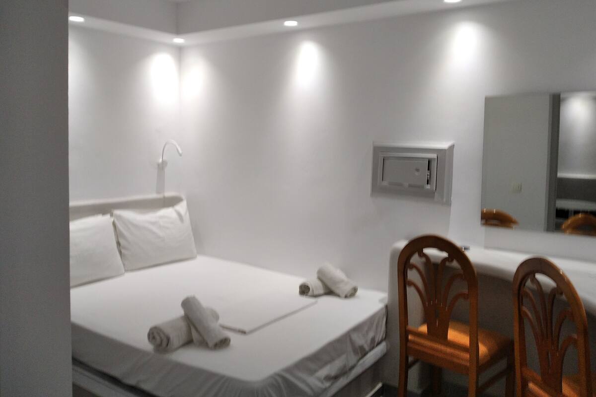 A hotel room with a bed, a desk, a chair, a mirror, and an air conditioning unit.