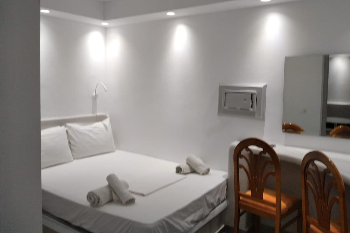 A hotel room with a bed, a desk, a chair, a mirror, and an air conditioning unit.