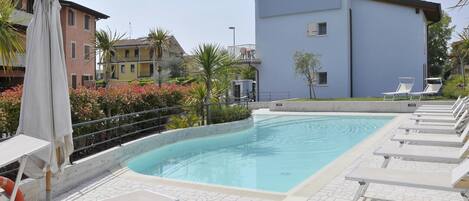 Una piscina con sillones y sombrillas, rodeada de edificios y follaje.
