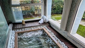 Un área de spa con un jacuzzi rodeado de guijarros blancos y suelo de baldosas, ventanas amplias con arcos y vistas a una zona residencial.