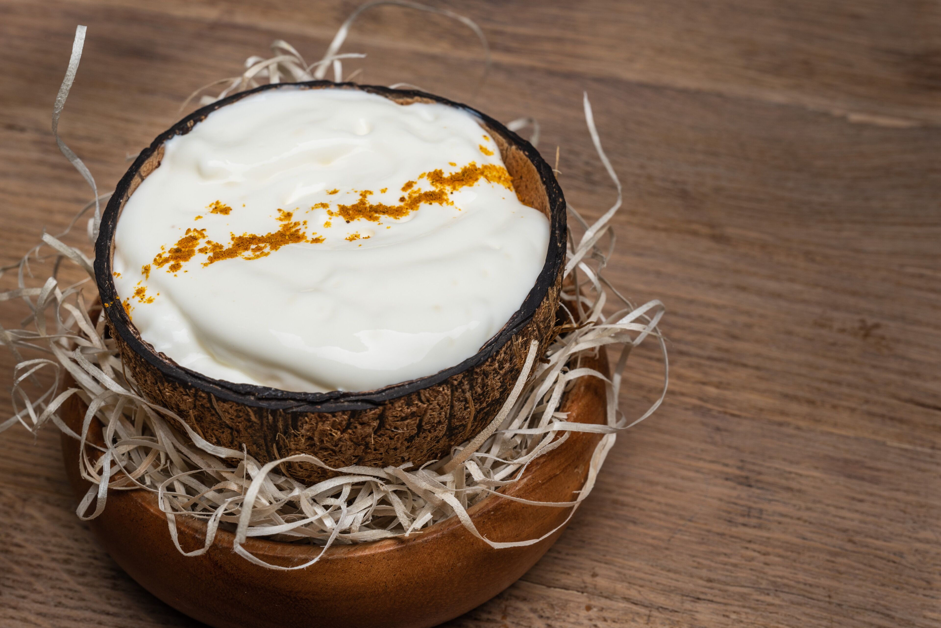 A coconut bowl filled with white substance, possibly yogurt, and sprinkled with a yellow spice, placed on a wooden surface.