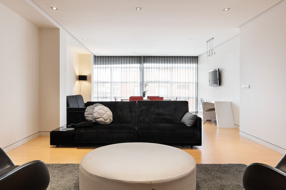 A modern living room with a black sofa, a round white ottoman, and a flat-screen TV mounted on the wall.