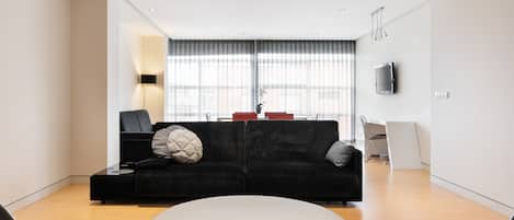 Een moderne woonkamer met een zwarte bank, een rond wit pouf en een flatscreen televisie aan de wand.