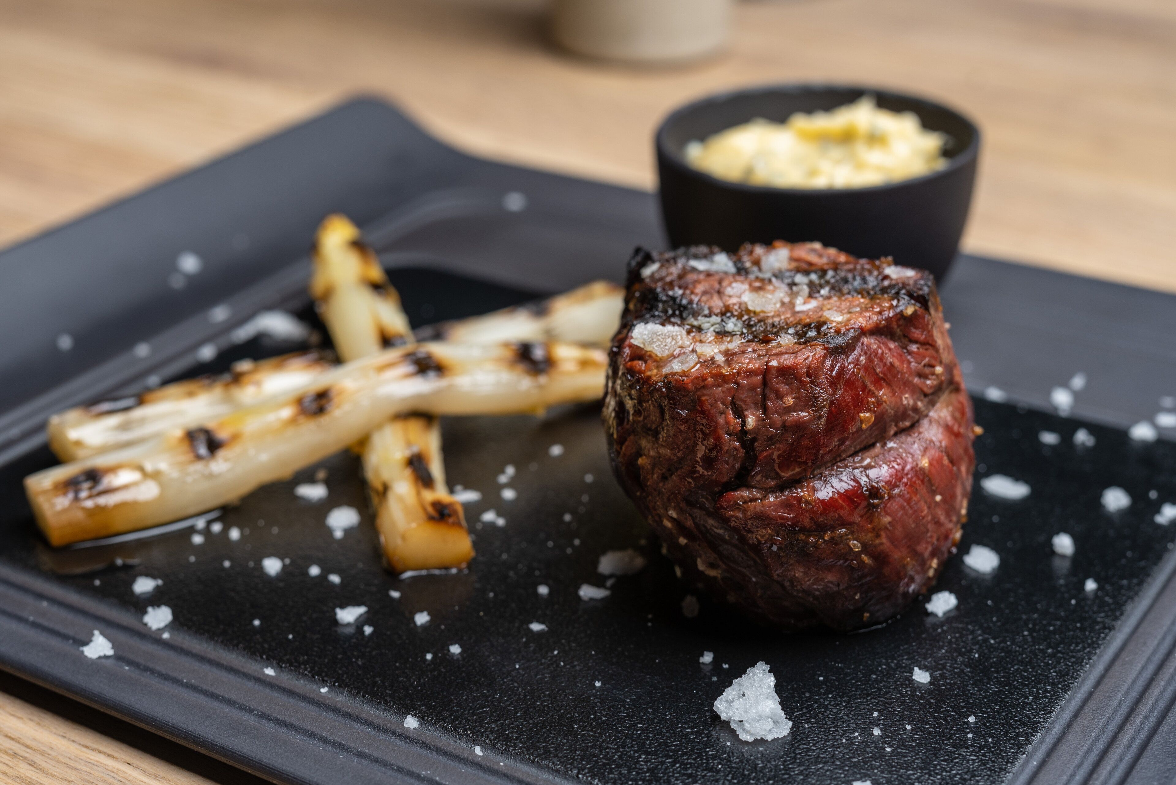 A steak with grilled vegetables on a black plate, accompanied by a small bowl of sauce.
