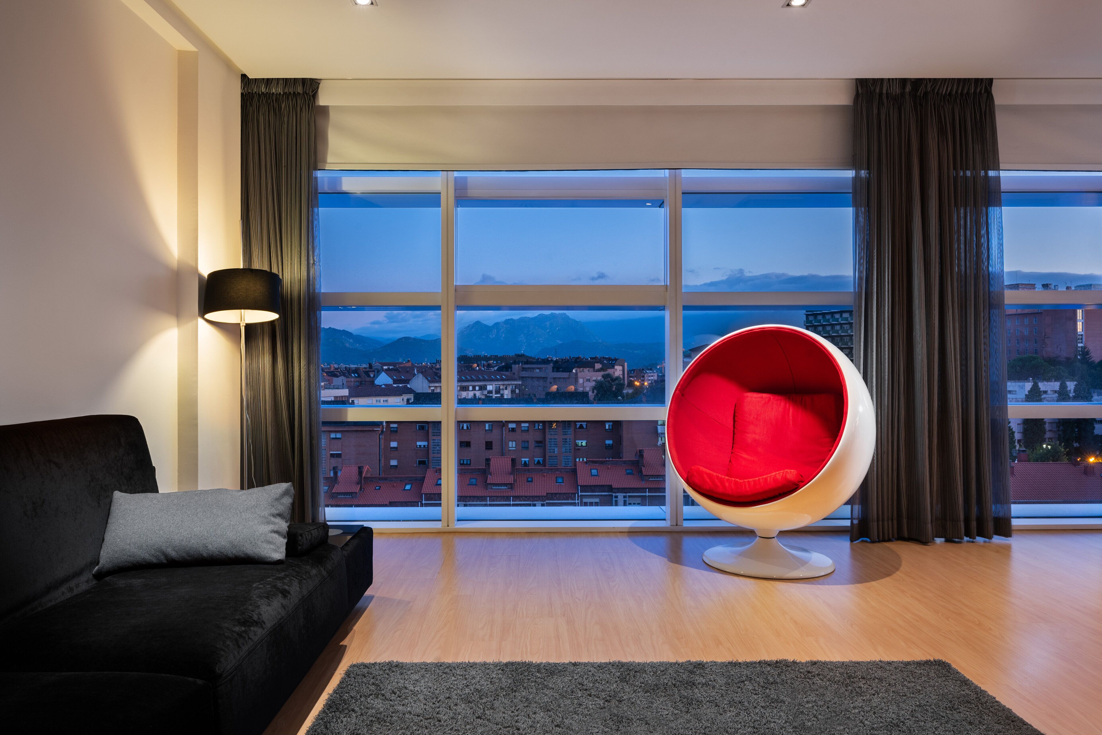 Un salon moderne avec un canapé noir, un fauteuil rouge et une grande fenêtre donnant sur la ville.