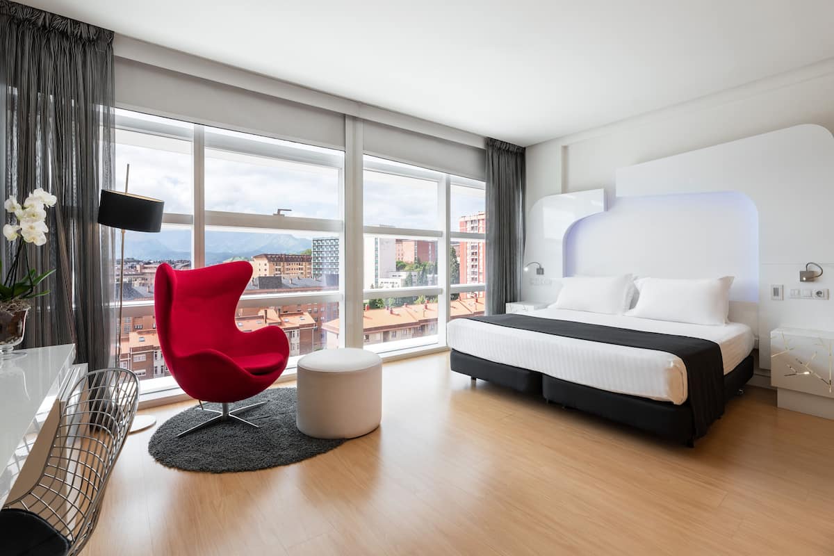 A modern hotel room with a large bed, a red chair, a flat-screen TV, and a view of the city.