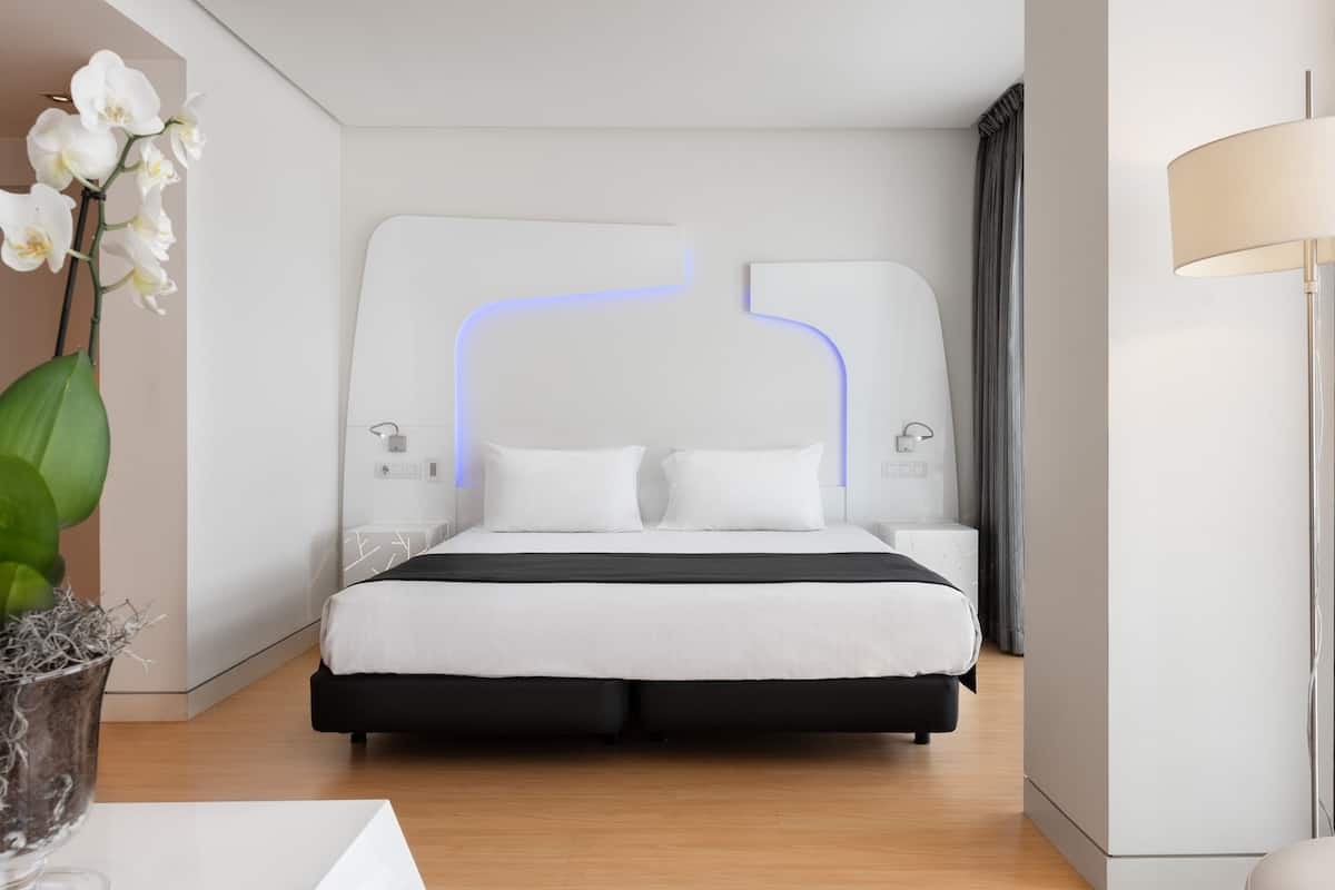 A modern hotel room with a bed, bedside tables, a lamp, and a vase with flowers.