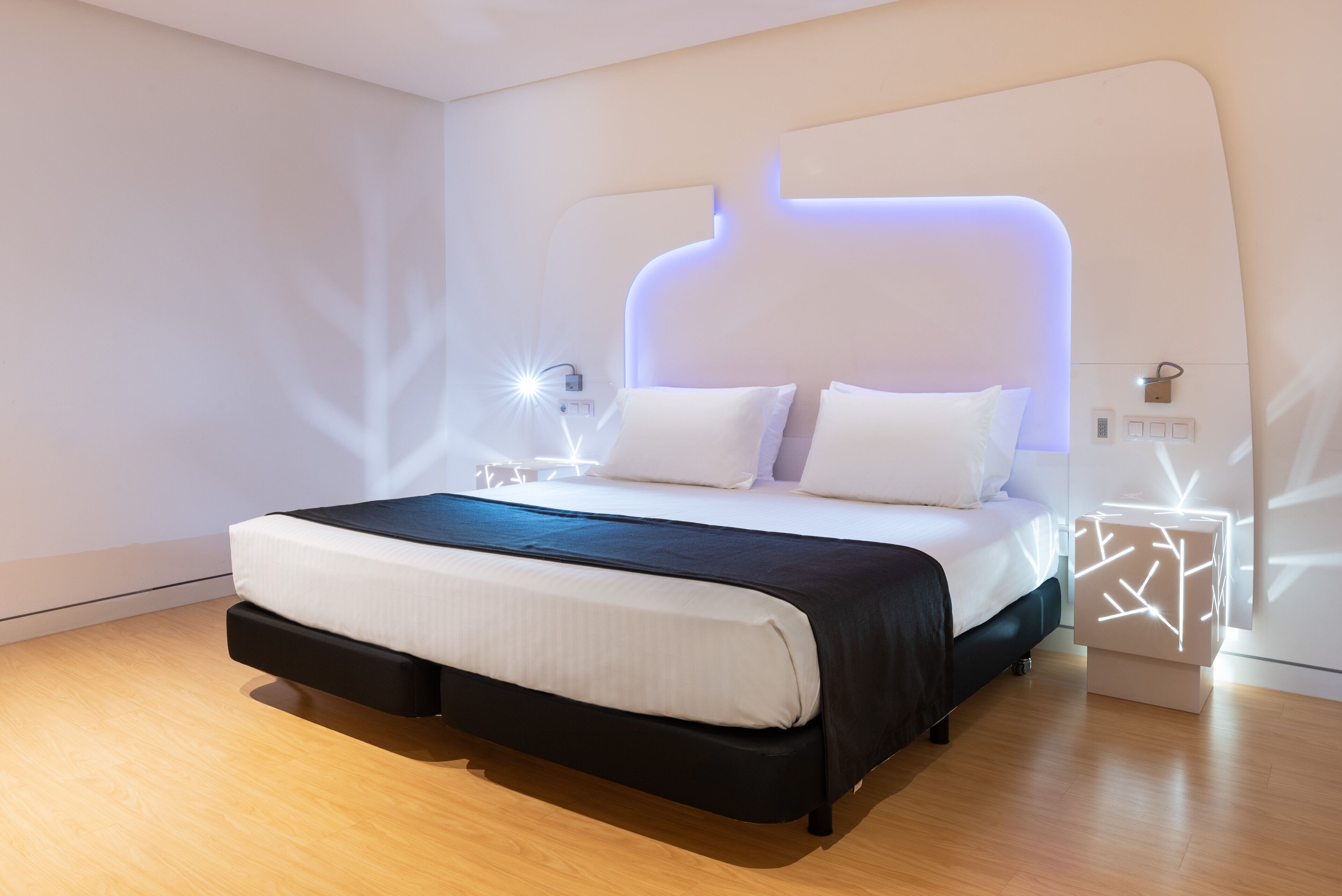 Un dormitorio moderno con una cama grande, cabecera con iluminación integrada, mesita de noche con lámpara y suelo de madera.