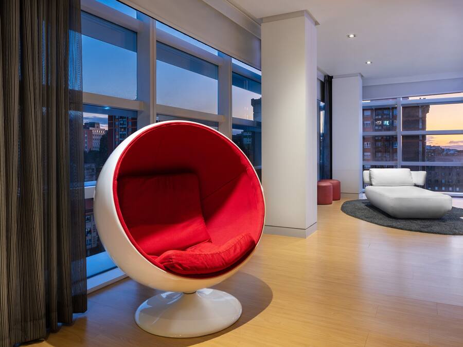 Een modern interieur met een rode eierstoel, een witte bank en een uitzicht op gebouwen door grote ramen.