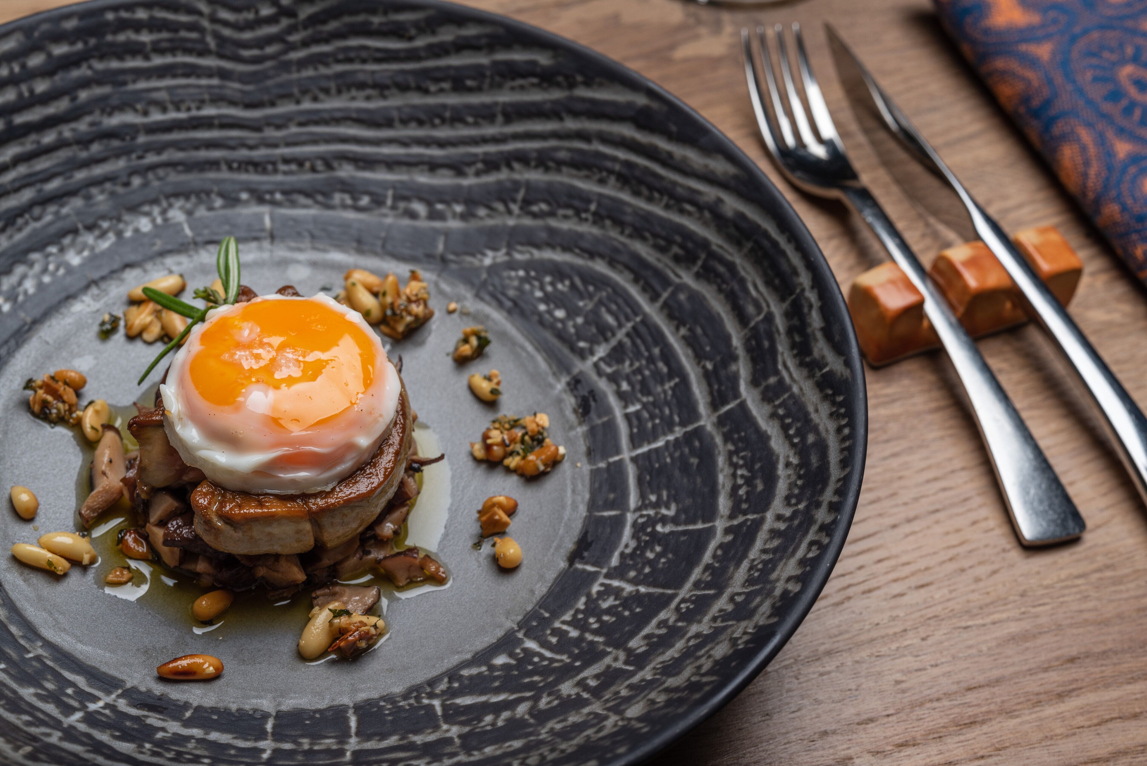 A plate with a poached egg on top of a stack of meat and vegetables, accompanied by nuts and a sprig of rosemary.