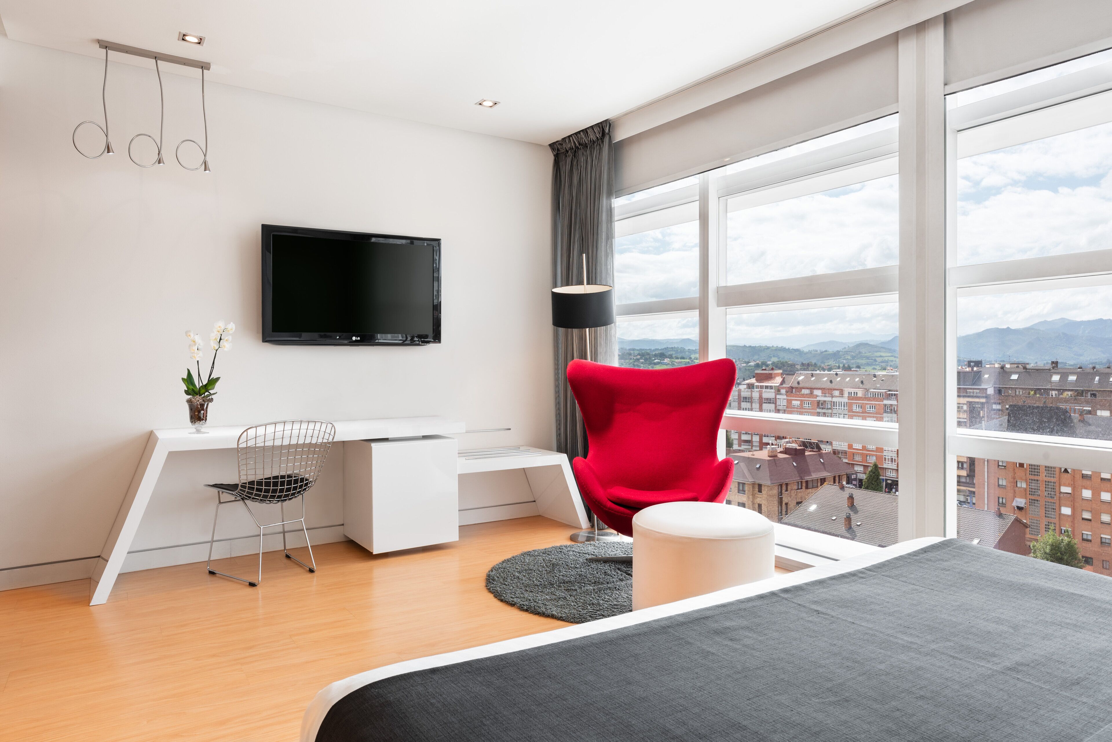 Ett modernt hotellrum med ett stort fönster, en platt-TV, en röd stol, ett skrivbord och en säng.