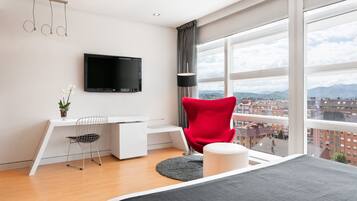 A modern hotel room with a large window, a flat-screen TV, a red chair, a desk, and a bed.