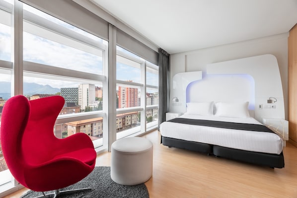 Una habitación de hotel moderna con una cama grande, una silla roja y vistas a la ciudad a través de amplios ventanales.