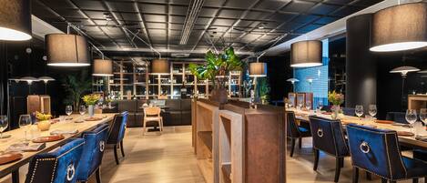A modern dining area with a long wooden table, blue upholstered chairs, and hanging pendant lights.