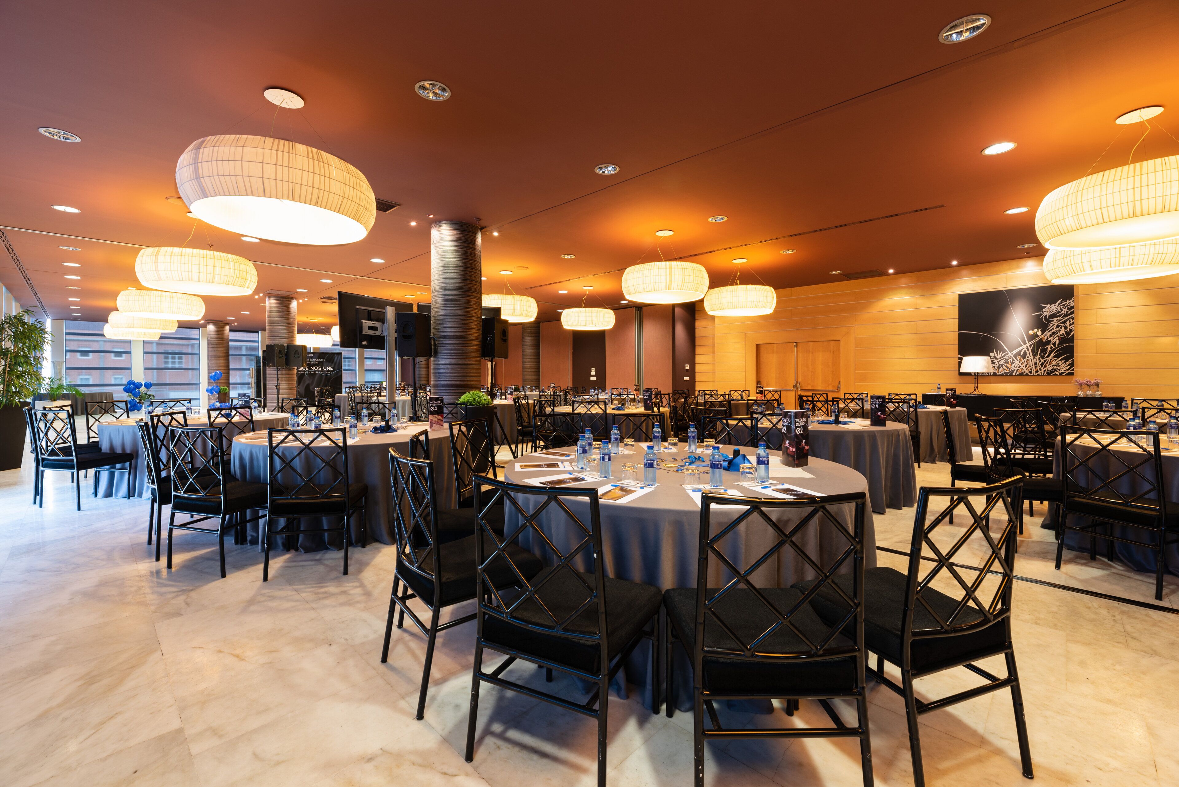 Un amplio salón de banquetes con mesas redondas y sillas negras, equipado con botellas de agua y menús.
