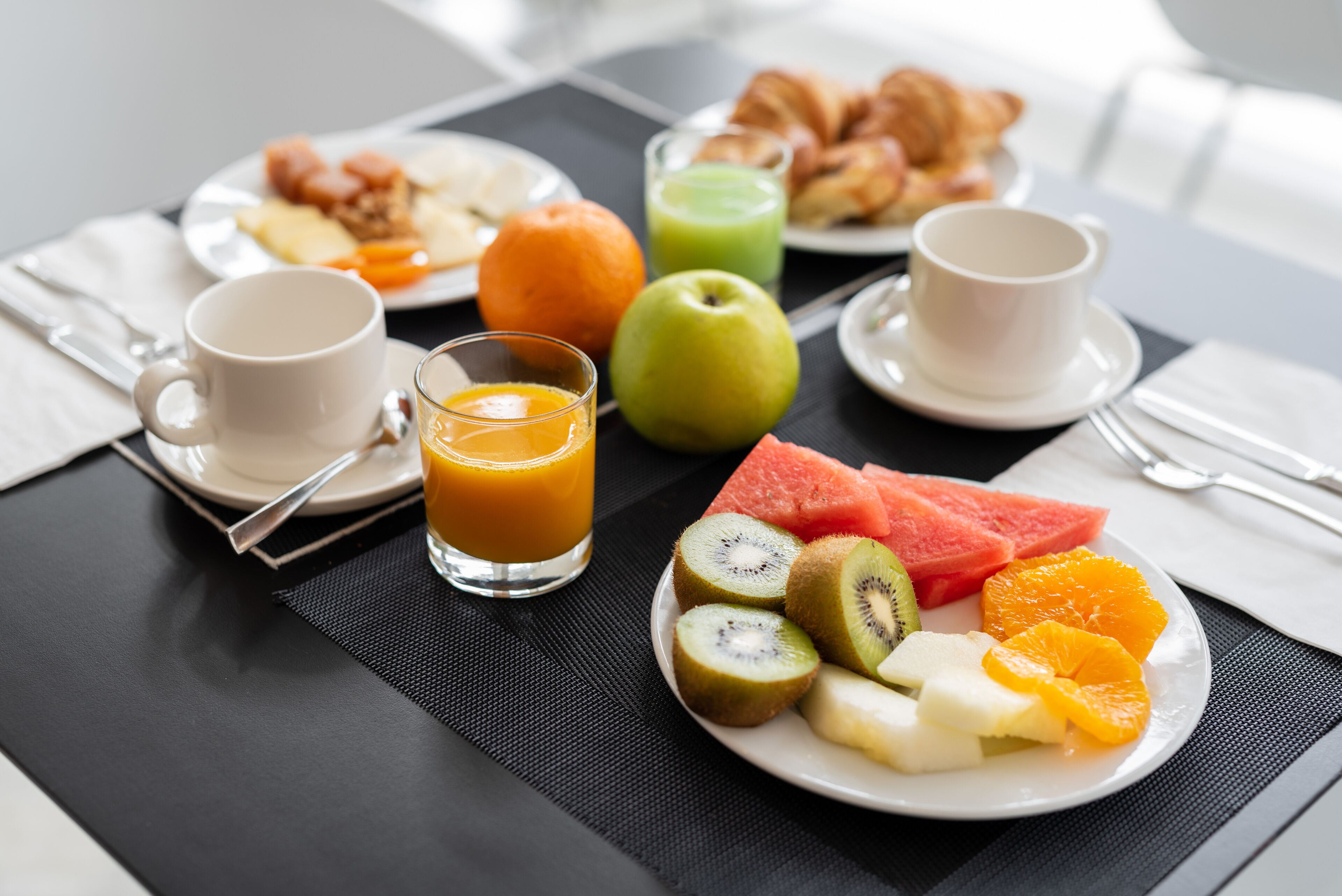 Mesa de café da manhã com pratos de frutas, croissants e suco.