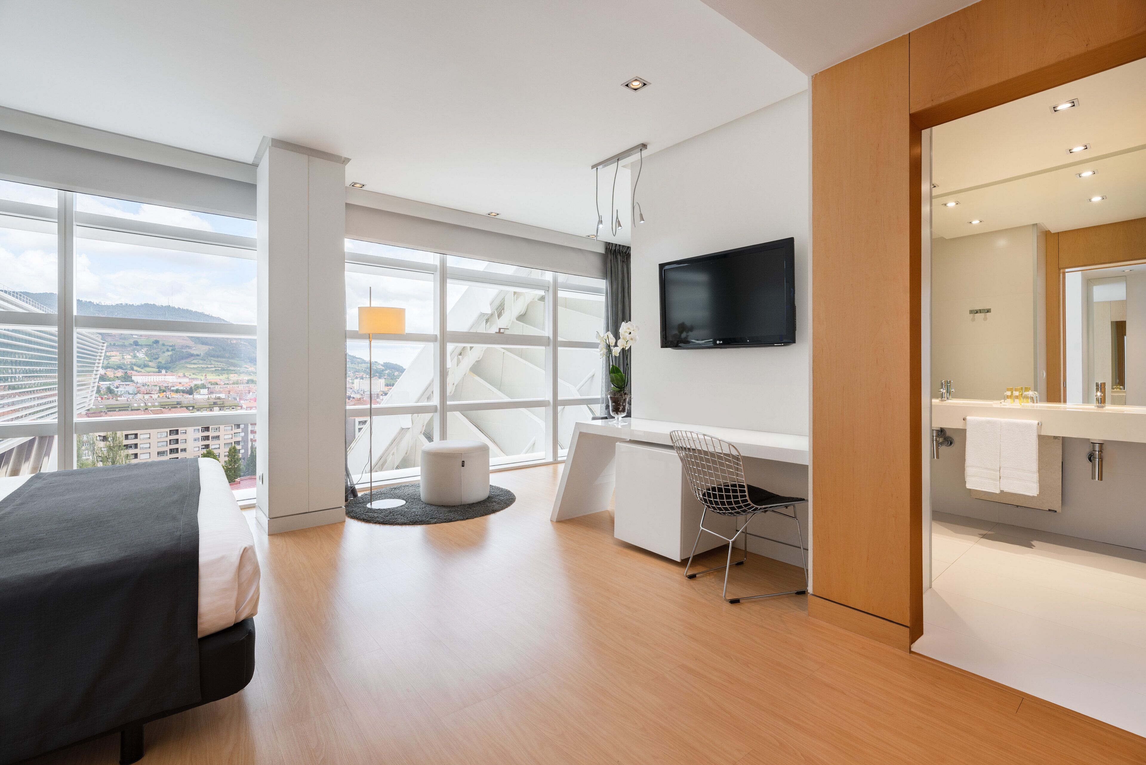 A modern hotel room with a large window, a bed, a desk, and a bathroom.
