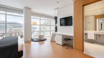 A modern hotel room with a large window, a bed, a desk, and a bathroom.