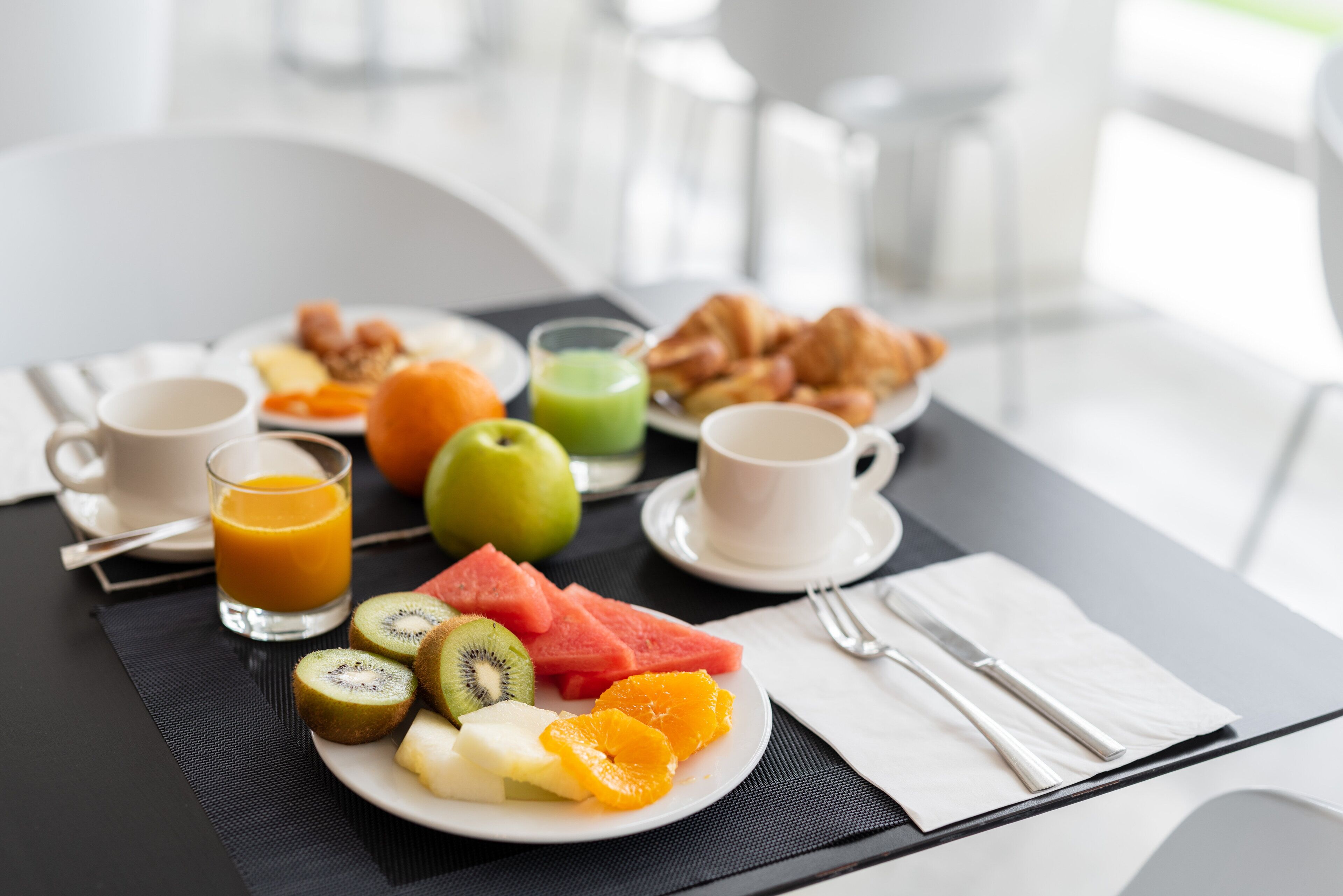 Un desayuno con un plato de fruta, cruasanes y un vaso de zumo de naranja sobre una mesa.