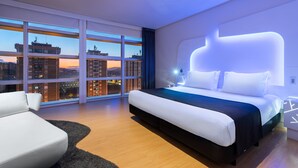 Habitació d'hotel moderna amb un llit gran, un sofà blanc i vista a la ciutat a través de grans finestres de sostre a terra.