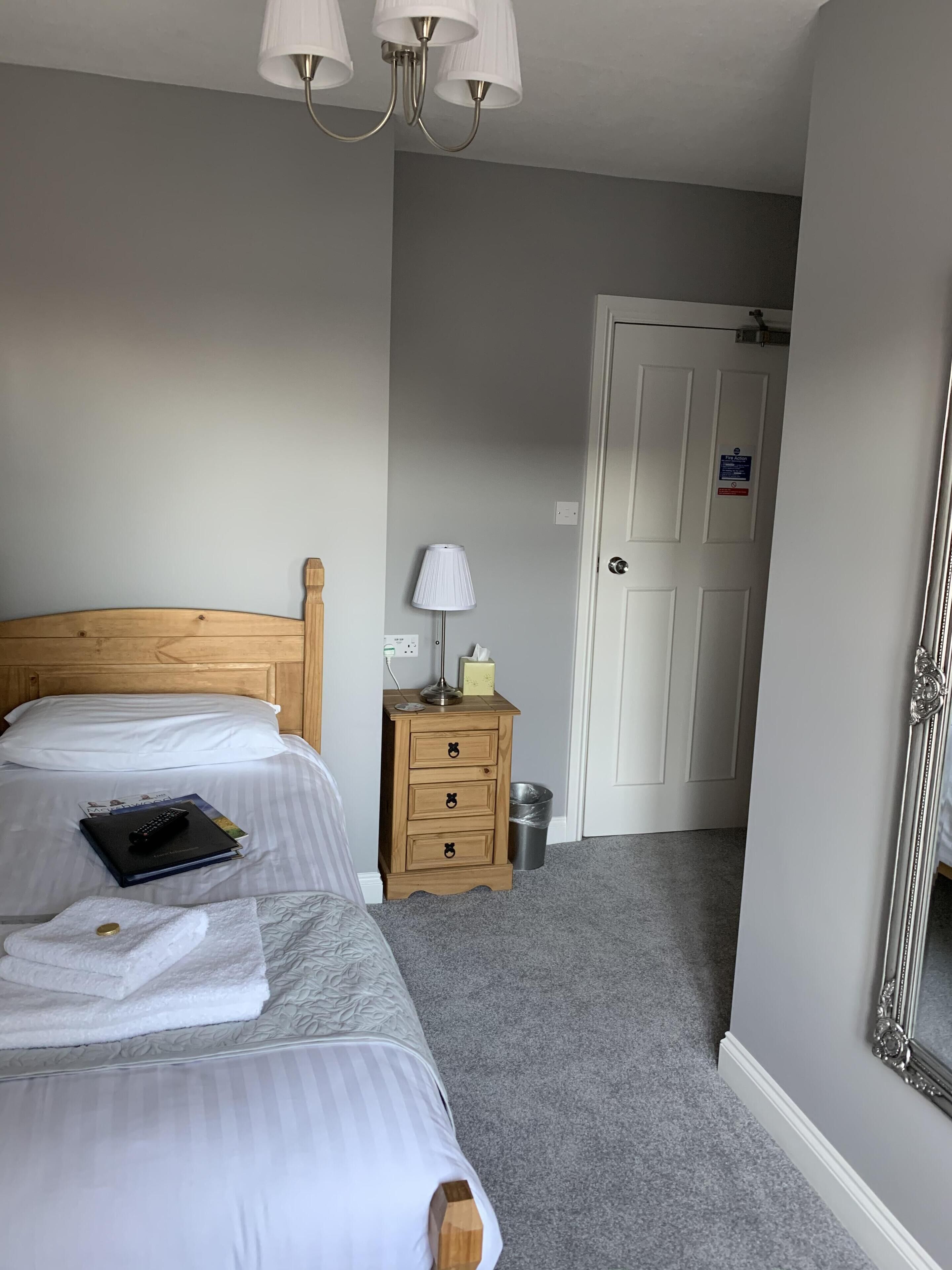 A hotel room with a wooden headboard, two beds, a bedside lamp, a nightstand, a trash bin, a door with a sign, and a mirror.
