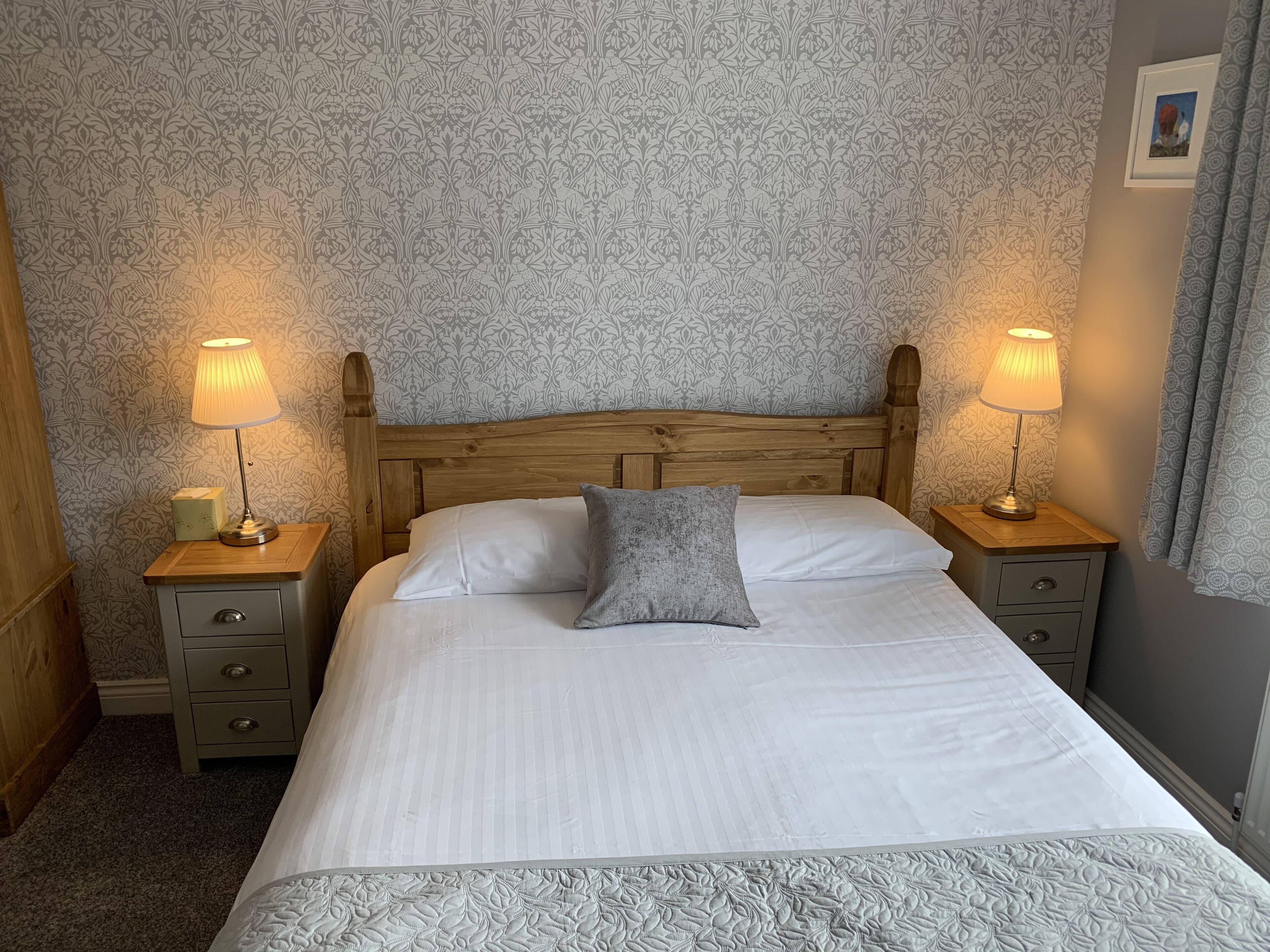 A neatly made bed with white linens and a grey pillow, flanked by two bedside tables with lamps, against a wallpapered wall.