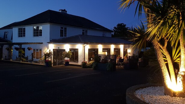 A white building with a dark roof, a covered patio area with seating, and a palm tree in the foreground. - Hensleigh House (Bridport)