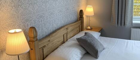 A neatly made bed with white linens, a wooden headboard, and a gray pillow. There is a bedside table with a lamp and a box, and a framed picture on the wall.