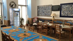 A dining area with a long table covered in a blue tablecloth, surrounded by wooden chairs. There is a television mounted on the wall and a decorative wall piece above the table. - Pouso Dos Anjos (Lisbon)