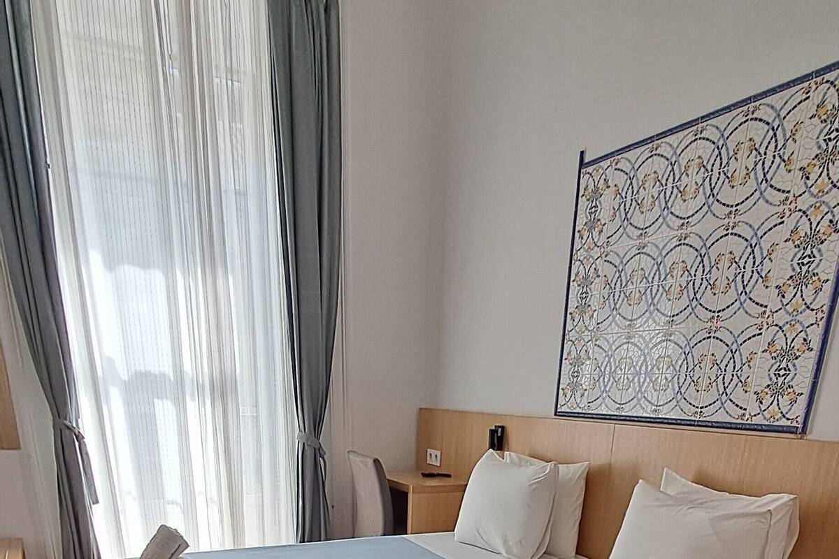 A double bed with two pillows and a blanket, a bedside table, a chair, and a large window with curtains.