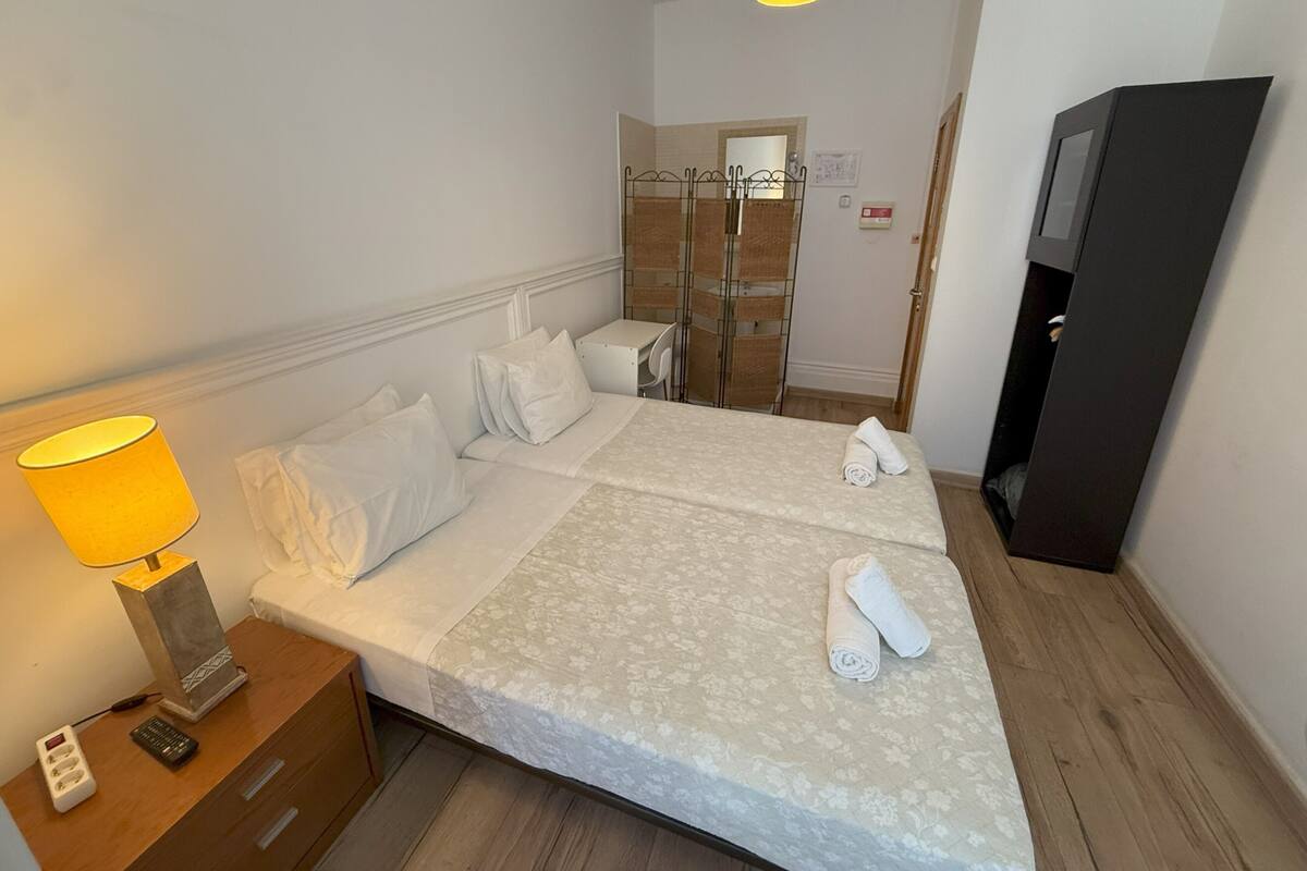 A double bed with a floral bedspread, two white pillows, and a rolled towel on the bed. There is a bedside table with a lamp and a remote control. A wardrobe is visible in the background.