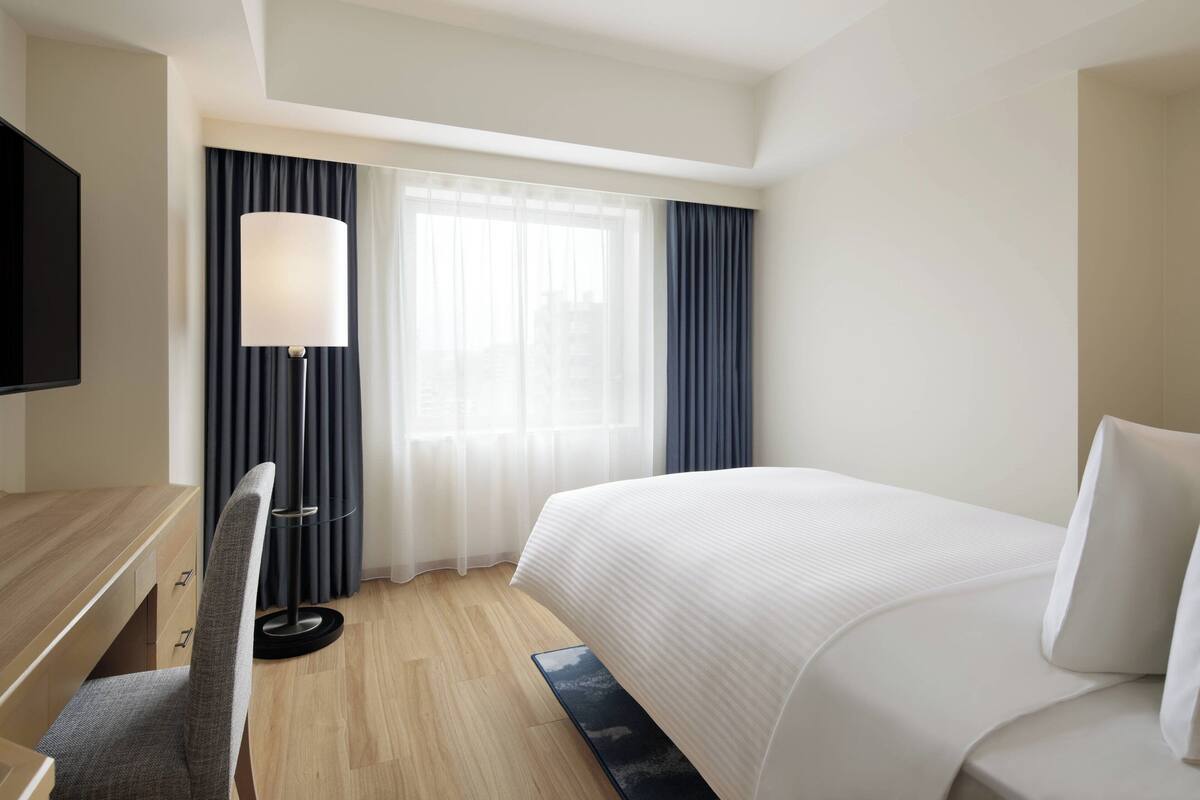 A hotel room with a bed, a desk, a chair, a TV, and a window with curtains.