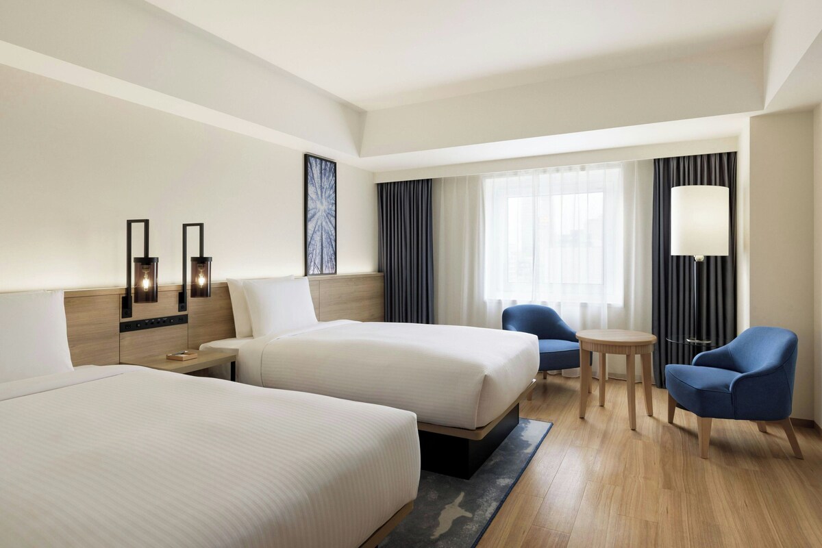 A hotel room with two beds, a desk, a chair, a small table, and a lamp.