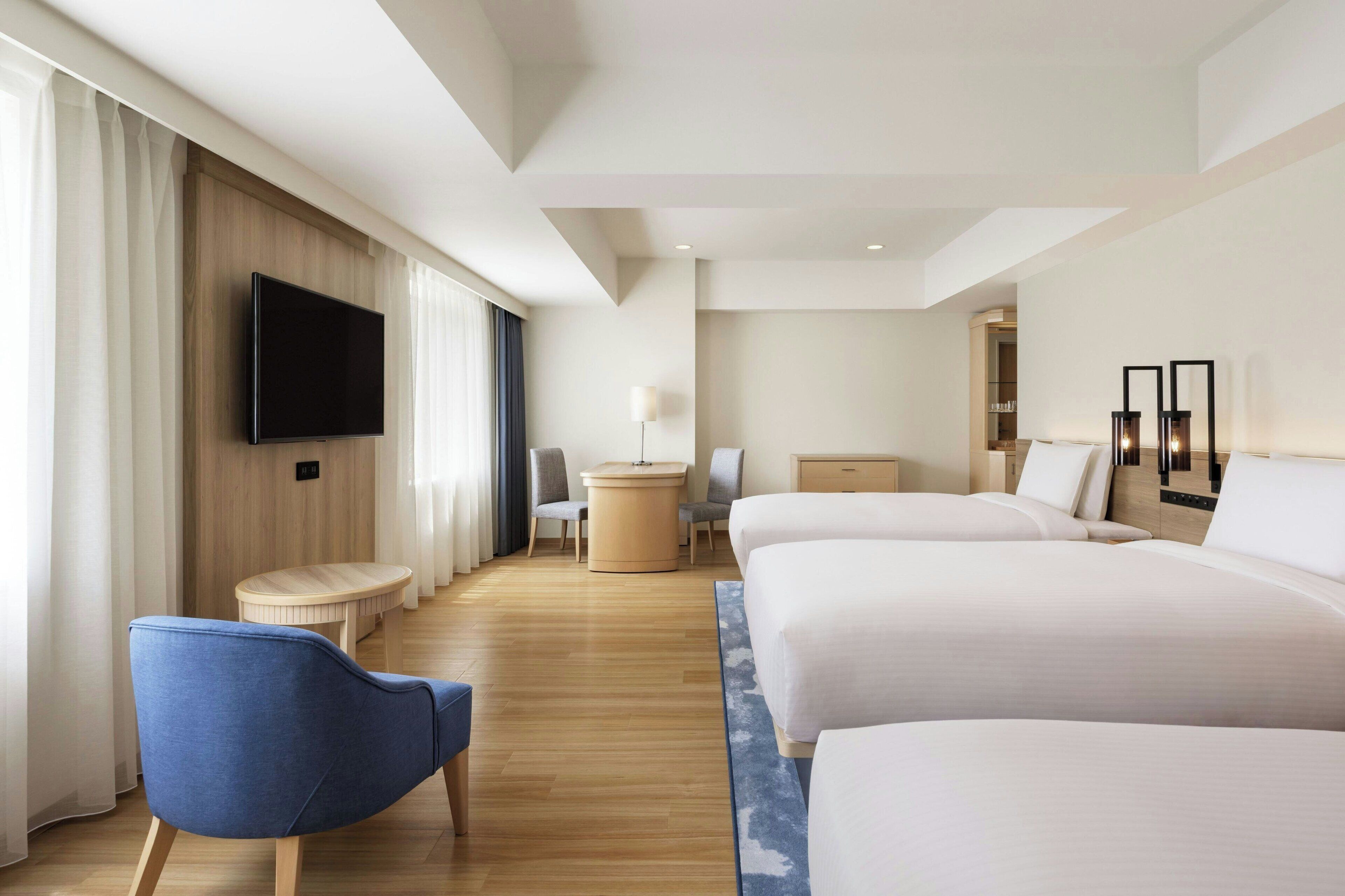 A modern hotel room with two large beds, a flat-screen TV mounted on the wall, a round table, and a blue chair.
