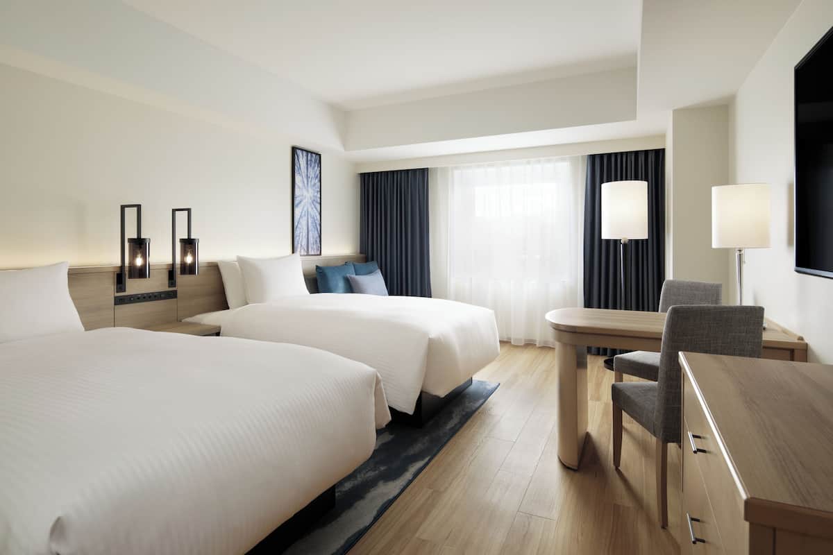 A hotel room with two beds, a desk, a chair, a TV, and a window with curtains.
