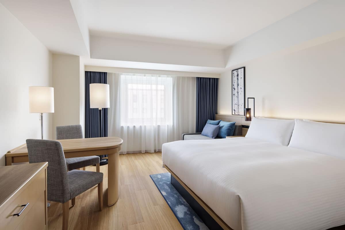 A hotel room with a large bed, a desk with a chair, a small table, and a window with curtains.