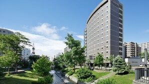 A modern hotel with a prominent name on the facade, surrounded by lush greenery and a small water feature. - Fairfield by Marriott Sapporo (Sapporo)