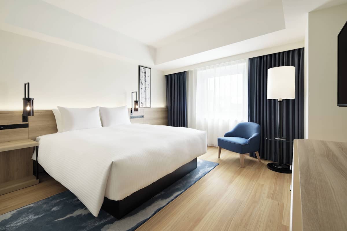 A modern hotel room with a large bed, a desk, a chair, and a lamp.
