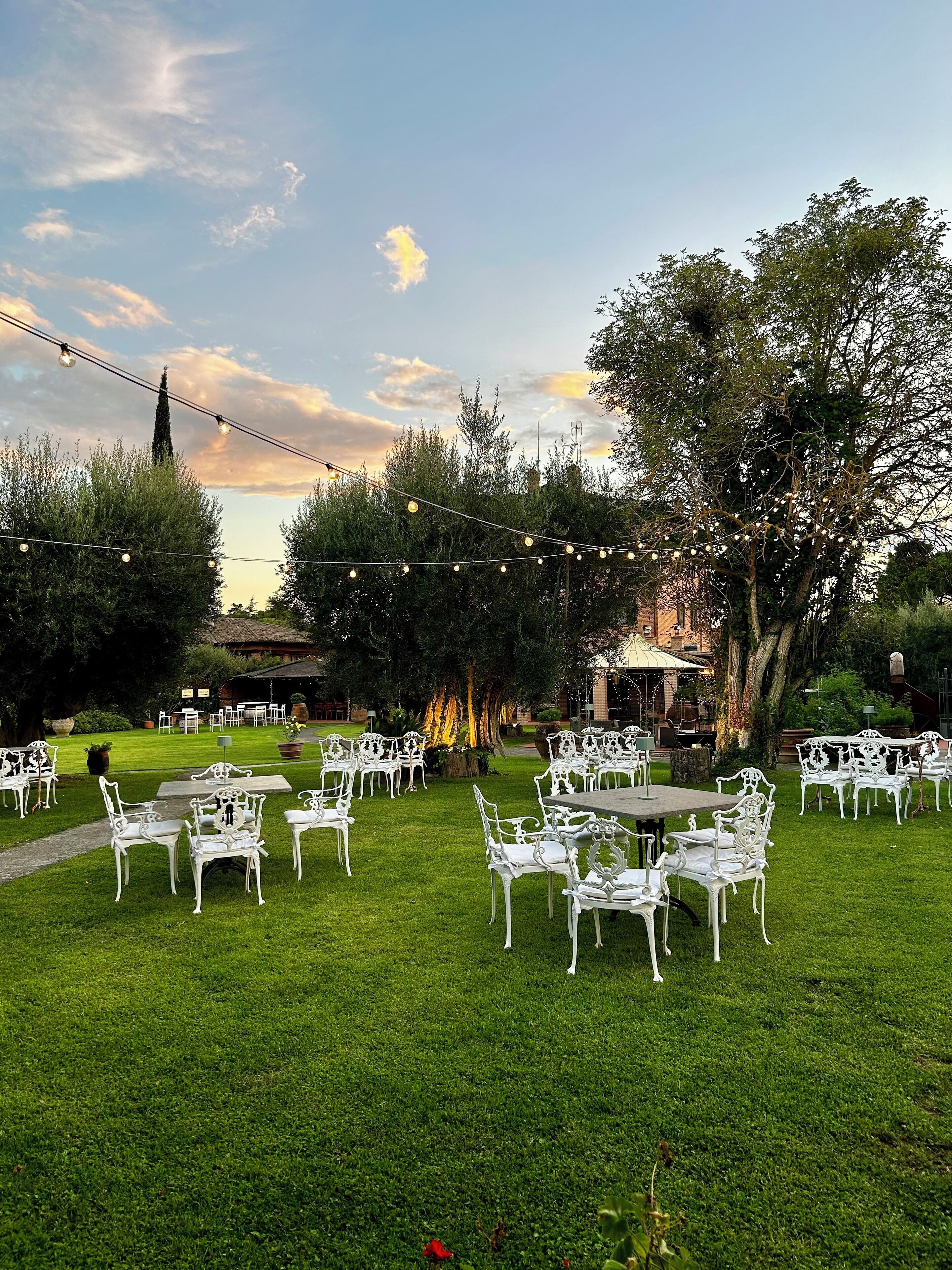 Een terras met witte metalen stoelen en tafels, omringd door groen en bomen, onder een heldere lucht.