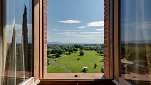 Vista dalla finestra su un muro di mattoni e telaio in legno, con una prospettiva su un paesaggio verde con alberi e cielo sereno.