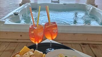 Un jacuzzi con vistas a una piscina y edificios, dos bebidas con pajitas y un plato con pan y ensalada.