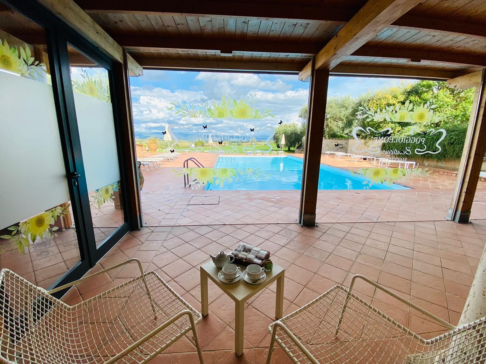 A covered patio with a pool, two metal chairs, a small table with a teapot and cup, and a view of the pool area.