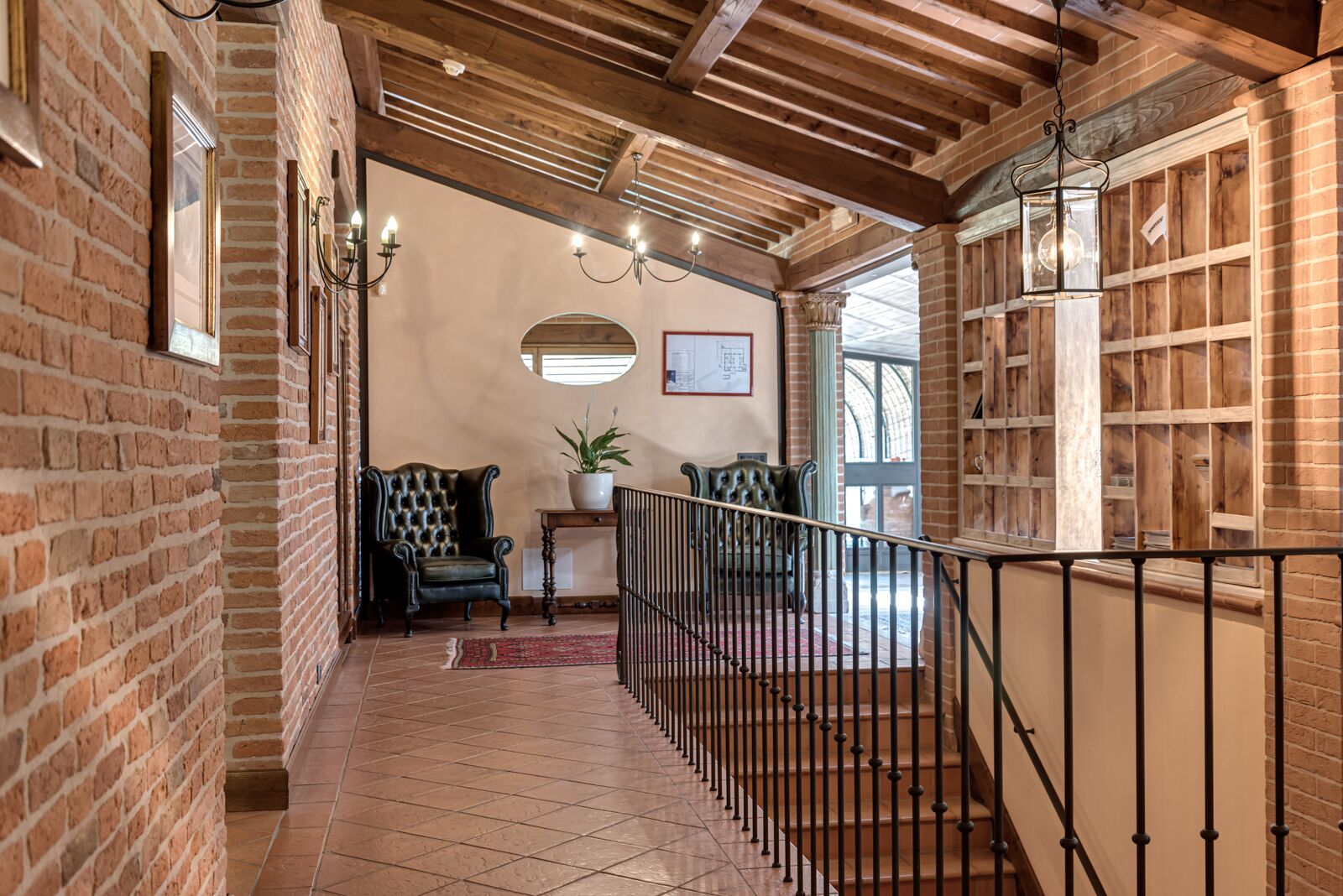 A hallway with brick walls, a wooden ceiling, and a tiled floor. There are two chairs, a small table with a plant, and a railing along the stairs.
