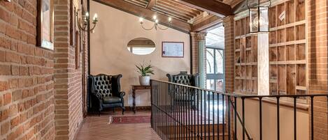 A hallway with brick walls, a wooden ceiling, and a tiled floor. There are two chairs, a small table with a plant, and a railing along the stairs.