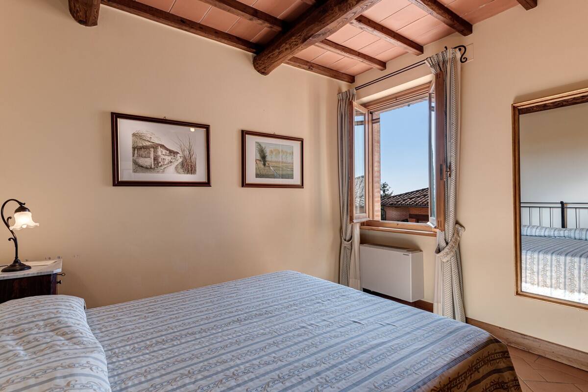Une chambre à coucher comprenant un lit, une table de chevet, une lampe, deux tableaux encadrés, une fenêtre avec des rideaux et un radiateur.