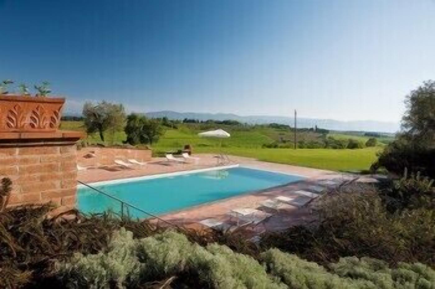A rectangular swimming pool with a tiled deck, surrounded by greenery and a clear sky.