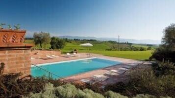 A rectangular swimming pool with a tiled deck, surrounded by greenery and a clear sky.
